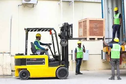 Forklift operator maneuvering a yellow forklift to lift a wooden crate, with workers in safety vests overseeing the loading process, illustrating efficient material handling in Australian industries.