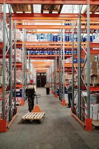 Warehouse interior with shelving units stacked with boxes, workers maneuvering pallet jack, emphasizing forklift accessory usage for efficiency and safety.