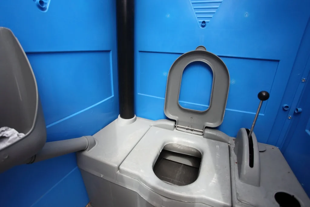 Interior view of a portable toilet featuring a gray toilet seat, a waste tank, and a handwashing station, set against a blue plastic backdrop, relevant to construction site sanitation and compliance.