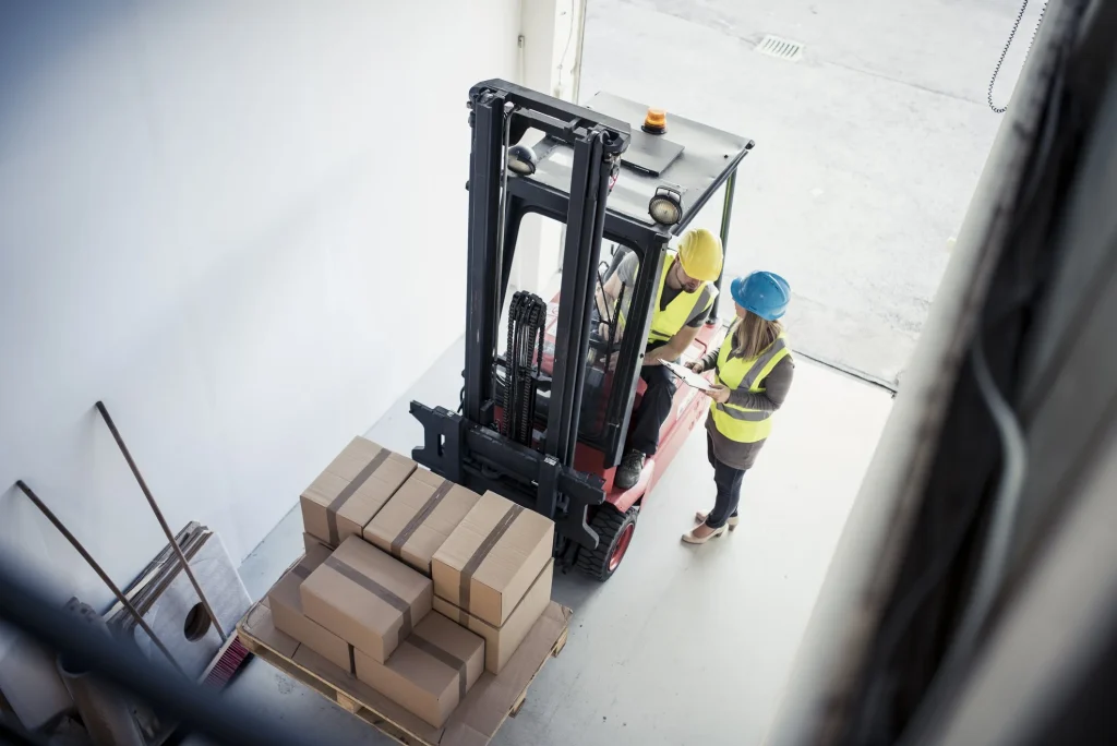 Workers in safety gear discussing forklift operation near stacked cardboard boxes in a warehouse setting, emphasizing safe and efficient material handling.