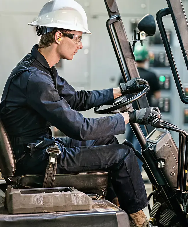 Worker operating a forklift in a warehouse setting, wearing a hard hat and safety glasses, focused on maneuvering equipment.