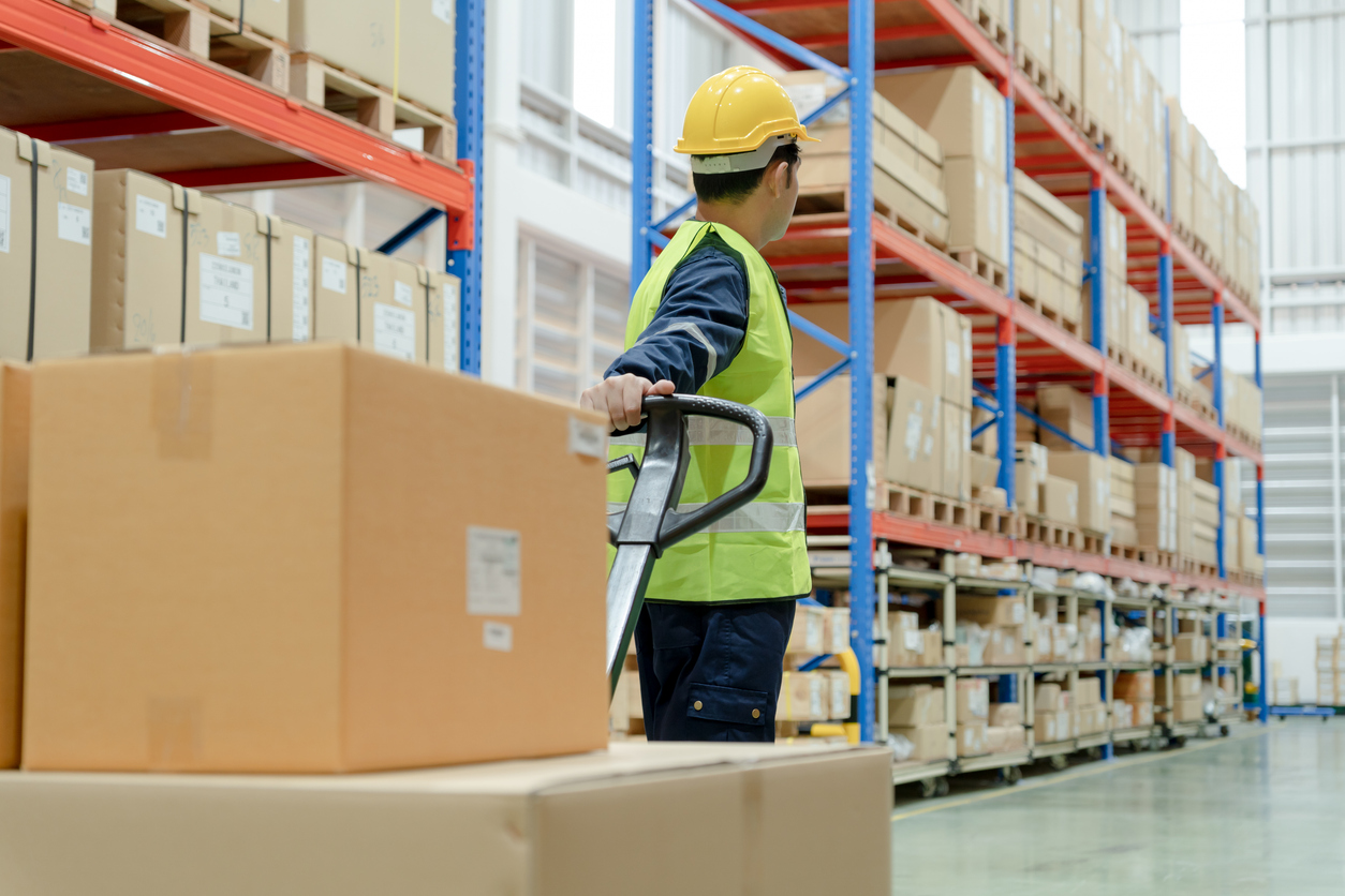 Warehouse worker operating hand pallet jack among stacked cardboard boxes in logistics facility, emphasizing efficiency in material handling.